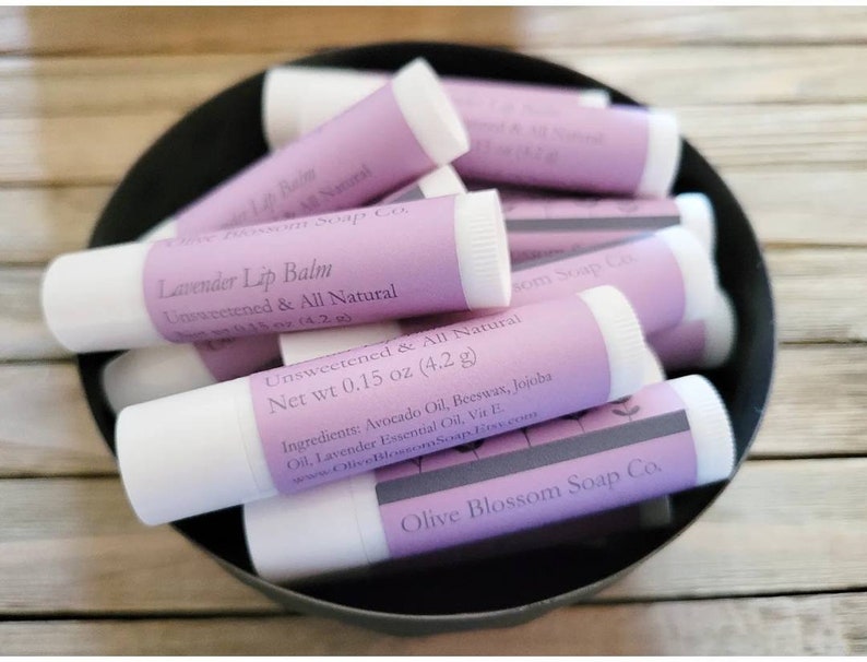 May include: A selection of lavender lip balm tubes in a black bowl. Each tube features a white base and a light purple label with the text "Lavender Lip Balm, Unsweetened & All Natural". The net weight is 4.2 grams.