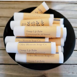 May include: A black bowl filled with several white lip balm tubes. Each tube has a light orange label with the text "Orange Lip Balm, Unsweetened & All Natural, Net wt 4.2 g" and "Olive Blossom Soap Co."