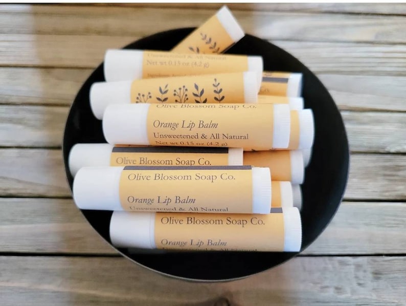May include: A black bowl filled with several white lip balm tubes. Each tube has a light orange label with the text "Orange Lip Balm, Unsweetened & All Natural, Net wt 4.2 g" and "Olive Blossom Soap Co."