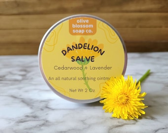 May include: A round silver tin of Dandelion Salve with a yellow label featuring the text olive blossom soap co., Dandelion Salve, Cedarwood + Lavender, An all natural soothing ointment, and Net Wt 2 Oz, accompanied by a bright yellow dandelion flower.