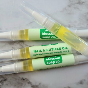 May include: Four clear nail and cuticle oil pens with white applicators and caps. Each pen has a light green label with the text "NAIL & CUTICLE OIL" and "Olive Blossom Soap Co.". The oil inside is yellow.