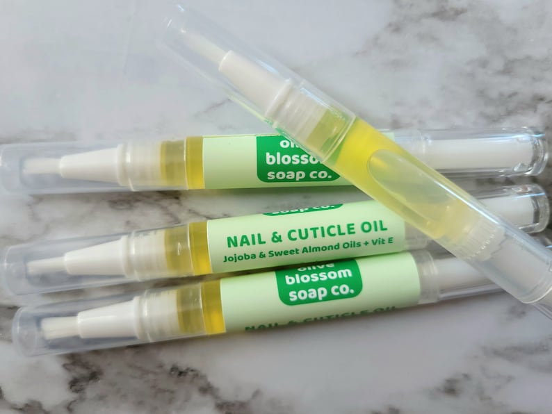 May include: Four clear nail and cuticle oil pens with white applicators and caps. Each pen has a light green label with the text "NAIL & CUTICLE OIL" and "Olive Blossom Soap Co.". The oil inside is yellow.
