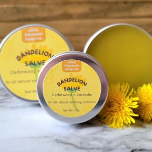 Puede incluir: Dos latas redondas de metal plateado que contienen ung&uuml;ento de diente de le&oacute;n con etiquetas amarillas. Las etiquetas muestran el texto Olive Blossom Soap Co., Dandelion Salve, Cedarwood + Lavender. Dos flores de diente de le&oacute;n amarillo brillante.