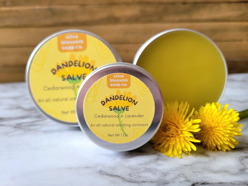 Puede incluir: Dos latas redondas de metal plateado que contienen ung&uuml;ento de diente de le&oacute;n con etiquetas amarillas. Las etiquetas muestran el texto Olive Blossom Soap Co., Dandelion Salve, Cedarwood + Lavender. Dos flores de diente de le&oacute;n amarillo brillante.