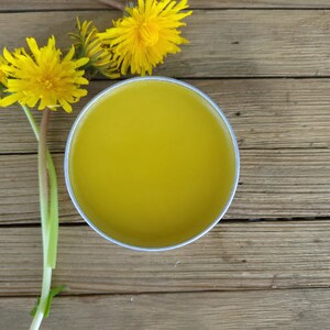 Puede incluir: Una lata redonda de metal plateado llena de un b&aacute;lsamo suave de color amarillo, colocada junto a tres flores de diente de le&oacute;n de color amarillo brillante con tallos verdes. El recipiente est&aacute; abierto, revelando la sustancia cerosa s&oacute;lida.