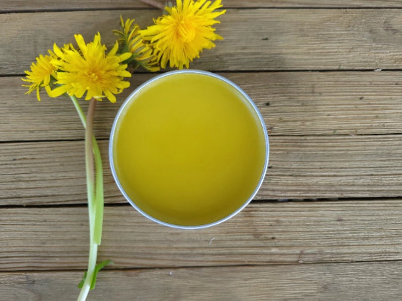Puede incluir: Una lata redonda de metal plateado llena de un b&aacute;lsamo suave de color amarillo, colocada junto a tres flores de diente de le&oacute;n de color amarillo brillante con tallos verdes. El recipiente est&aacute; abierto, revelando la sustancia cerosa s&oacute;lida.
