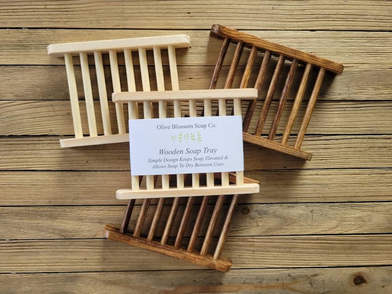 May include: Three wooden soap trays in varying shades of brown and natural wood tones are displayed on a wooden surface. The trays have a slatted design. A card reads "Olive Blossom Soap Co. Wooden Soap Tray".