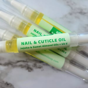 May include: Several nail and cuticle oil pens are displayed. Each pen has a clear plastic tube with a white applicator tip and a green label that reads "NAIL & CUTICLE OIL" with the ingredients listed. The oil inside is yellow.