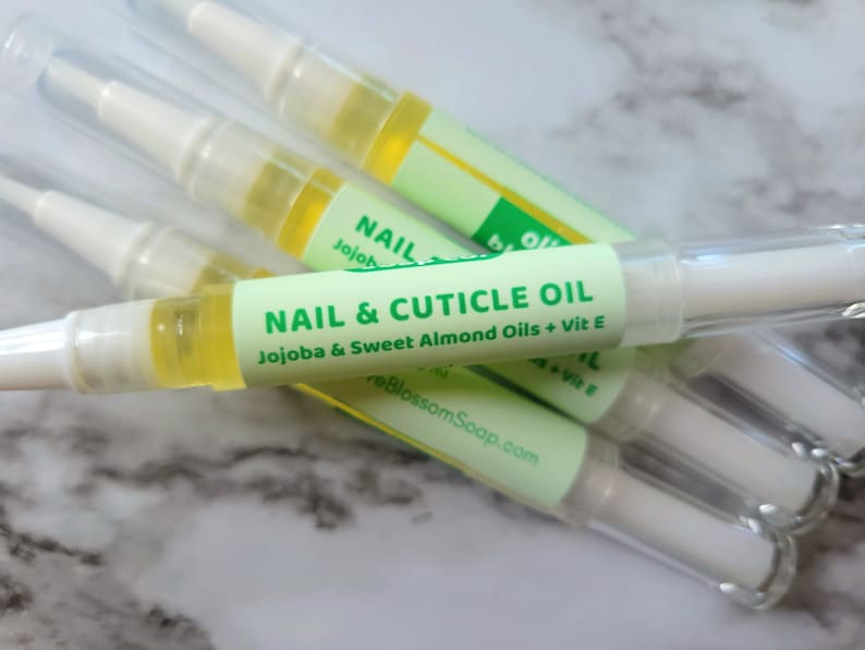 May include: Several nail and cuticle oil pens are displayed. Each pen has a clear plastic tube with a white applicator tip and a green label that reads "NAIL & CUTICLE OIL" with the ingredients listed. The oil inside is yellow.