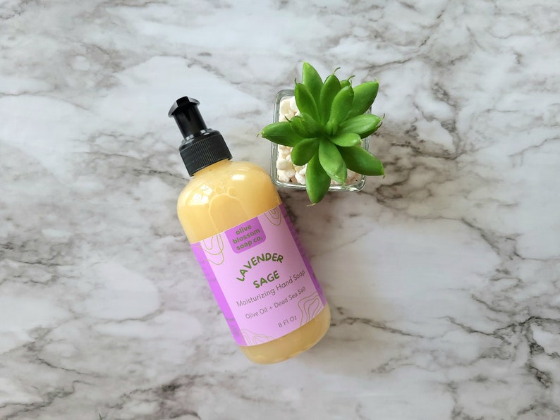 May include: A clear plastic bottle of yellow liquid hand soap with a black pump dispenser. The label is purple with white text reading Olive Blossom Soap Co., Lavender Sage, Moisturizing Hand Soap, Olive Oil + Dead Sea Salt, 8 Fl Oz.
