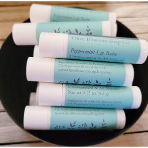 May include: Several white lip balm tubes with light blue labels are stacked in a black bowl. The labels state "Peppermint Lip Balm" and "Unsweetened & All Natural." The net weight is 4.2 g.