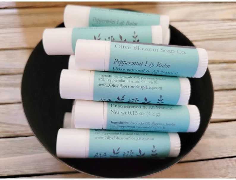 May include: Several white lip balm tubes with light blue labels are stacked in a black bowl. The labels state "Peppermint Lip Balm" and "Unsweetened & All Natural." The net weight is 4.2 g.