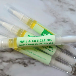 May include: Several nail and cuticle oil pens are displayed. Each pen has a clear plastic tube with a white applicator tip and a green label that reads "NAIL & CUTICLE OIL" with the ingredients listed. The oil is a light yellow color.