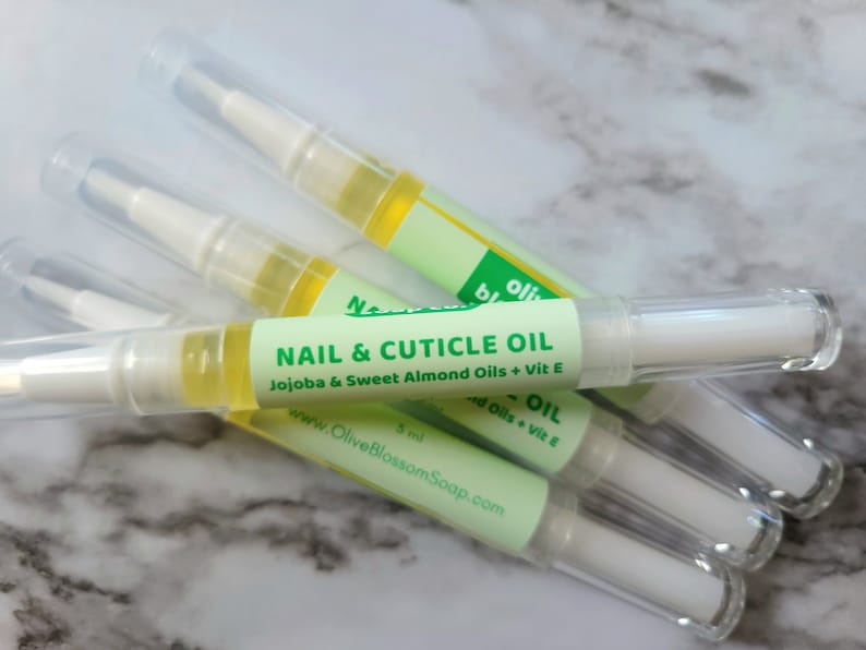 May include: Several nail and cuticle oil pens are displayed. Each pen has a clear plastic tube with a white applicator tip and a green label that reads "NAIL & CUTICLE OIL" with the ingredients listed. The oil is a light yellow color.