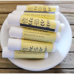 May include: Several white lip balm tubes with yellow labels are arranged in a white bowl. The labels display "Lemon Mint Lip Balm" and "Unsweetened & All Natural." The net weight is 4.2 grams.