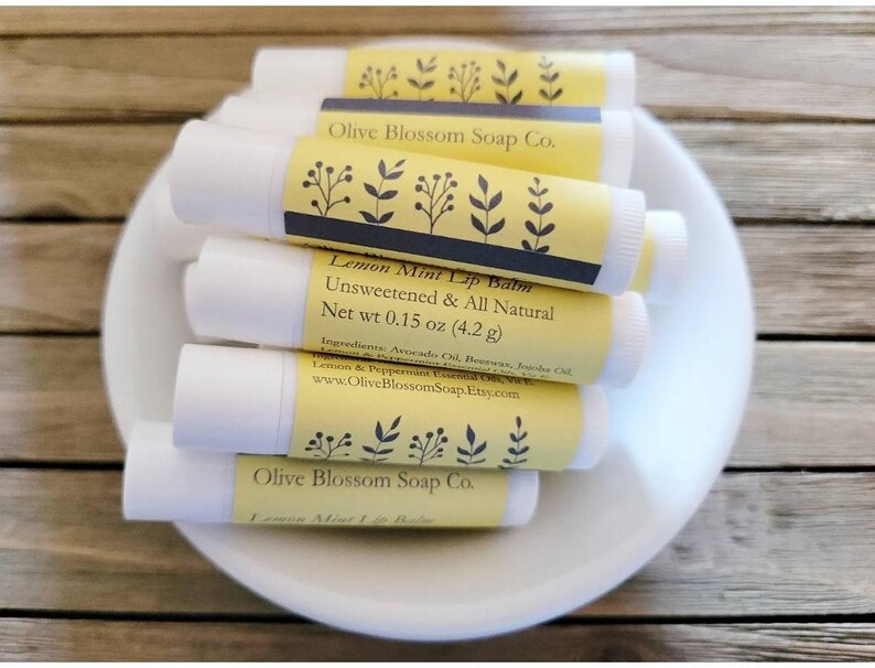 May include: Several white lip balm tubes with yellow labels are arranged in a white bowl. The labels display "Lemon Mint Lip Balm" and "Unsweetened & All Natural." The net weight is 4.2 grams.