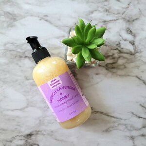May include: An 8 fluid ounce bottle of moisturizing hand soap with a black pump dispenser. The label is light purple with the text Olive Blossom Soap Co. French Lavender & Honey Moisturizing Hand Soap Olive Oil + Dead Sea Salt.