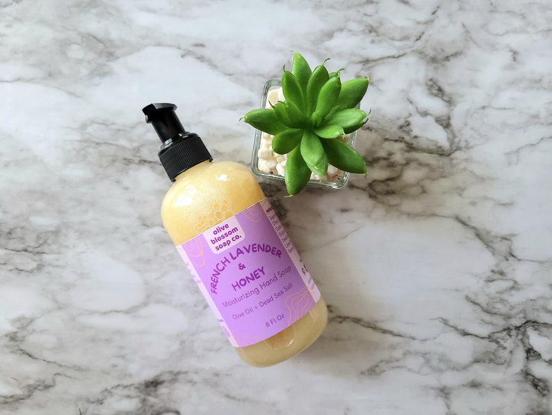 May include: An 8 fluid ounce bottle of moisturizing hand soap with a black pump dispenser. The label is light purple with the text Olive Blossom Soap Co. French Lavender & Honey Moisturizing Hand Soap Olive Oil + Dead Sea Salt.