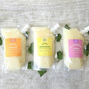 May include: Three 8 fl oz clear plastic pouches of moisturizing hand soap from Olive Blossom Soap Co. The labels are orange, yellow, and pink, featuring the scents Orange Blossom, Citrus Lemongrass, and Black Raspberry Vanilla with Olive Oil and Dead Sea Salt.