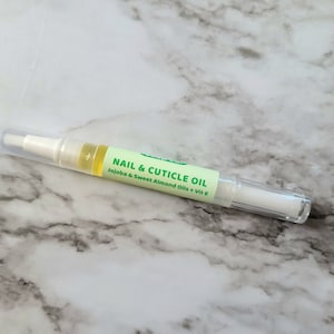 May include: A clear nail and cuticle oil pen with a white applicator tip and a clear cap. The pen has a green label with the text "NAIL & CUTICLE OIL" and the ingredients "Jojoba & Sweet Almond Oils + Vit E".
