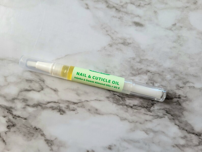 May include: A clear nail and cuticle oil pen with a white applicator tip and a clear cap. The pen has a green label with the text "NAIL & CUTICLE OIL" and the ingredients "Jojoba & Sweet Almond Oils + Vit E".
