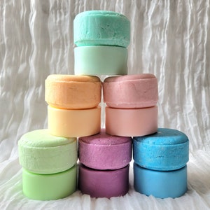 May include: A pyramid stack of nine round solid shampoo bars in various pastel colors including mint green, peach, light pink, pale yellow, lime green, lavender, and bright blue. Each bar has a smooth, cylindrical shape with flat top and bottom surfaces.