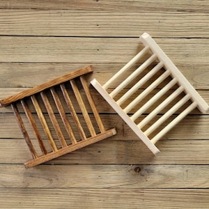 May include: Two wooden soap dishes. One is stained a dark brown, the other is natural wood. Each has a rectangular frame with parallel wooden slats. The dishes are designed to allow soap to drain and dry.