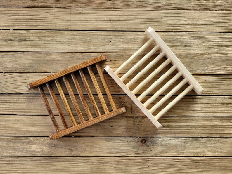 May include: Two wooden soap dishes. One is stained a dark brown, the other is natural wood. Each has a rectangular frame with parallel wooden slats. The dishes are designed to allow soap to drain and dry.