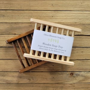 May include: Two wooden soap trays, one light and one dark, are displayed on a wooden surface. The light-colored tray has a white label that reads "Olive Blossom Soap Co. Wooden Soap Tray". The design allows soap to dry between uses.