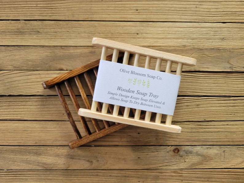 May include: Two wooden soap trays, one light and one dark, are displayed on a wooden surface. The light-colored tray has a white label that reads "Olive Blossom Soap Co. Wooden Soap Tray". The design allows soap to dry between uses.