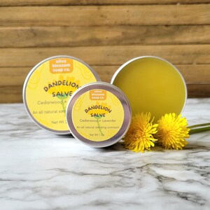Puede incluir: Dos latas redondas de metal con b&aacute;lsamo de diente de le&oacute;n con etiquetas amarillas que muestran el texto Olive Blossom Soap Co., Dandelion Salve, Cedarwood + Lavender. El b&aacute;lsamo es un ung&uuml;ento amarillo p&aacute;lido, acompa&ntilde;ado de dos flores amarillas.