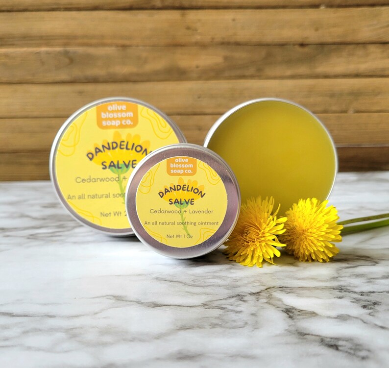Puede incluir: Dos latas redondas de metal con b&aacute;lsamo de diente de le&oacute;n con etiquetas amarillas que muestran el texto Olive Blossom Soap Co., Dandelion Salve, Cedarwood + Lavender. El b&aacute;lsamo es un ung&uuml;ento amarillo p&aacute;lido, acompa&ntilde;ado de dos flores amarillas.