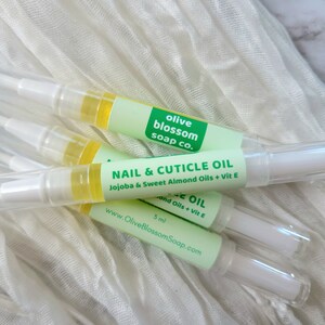 May include: Several nail and cuticle oil pens with clear tubes and white applicators are displayed. Each pen has a green label with the text "NAIL & CUTICLE OIL" and "Olive Blossom Soap Co.". The oil is yellow, and the pens are on a white cloth.