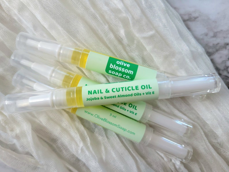 May include: Several nail and cuticle oil pens with clear tubes and white applicators are displayed. Each pen has a green label with the text "NAIL & CUTICLE OIL" and "Olive Blossom Soap Co.". The oil is yellow, and the pens are on a white cloth.