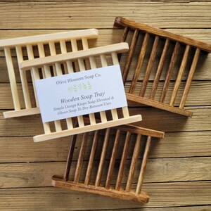 May include: Three wooden soap trays in varying shades of brown and natural wood. The trays have a slatted design with vertical bars. A white card with the text "Wooden Soap Tray" is visible on one tray. The text reads "Simple Design Keeps Soap Elevated & Allows Soap To Dry Between Uses."