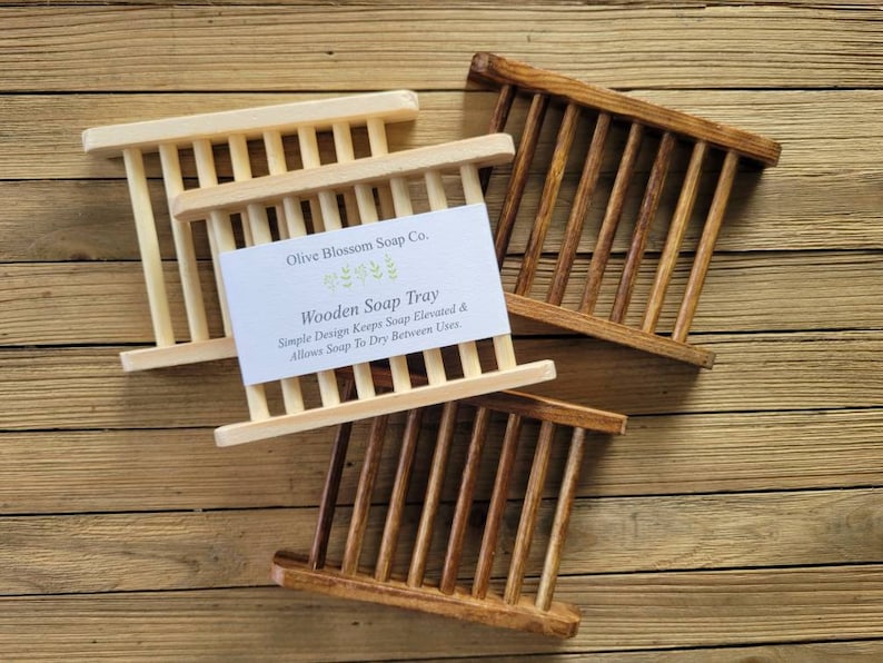 May include: Three wooden soap trays in varying shades of brown and natural wood. The trays have a slatted design with vertical bars. A white card with the text "Wooden Soap Tray" is visible on one tray. The text reads "Simple Design Keeps Soap Elevated & Allows Soap To Dry Between Uses."