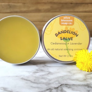 Puede incluir: Dos latas redondas de metal con b&aacute;lsamo de diente de le&oacute;n. Una lata est&aacute; abierta y muestra un ung&uuml;ento amarillo suave. La otra lata tiene una etiqueta amarilla que dice olive blossom soap co. DANDELION SALVE Cedarwood + Lavender Net Wt 57g.