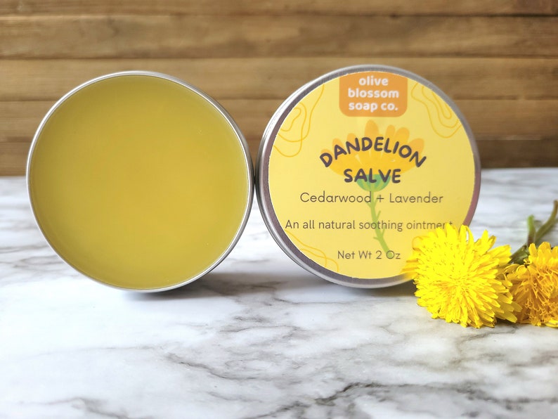 Puede incluir: Dos latas redondas de metal con b&aacute;lsamo de diente de le&oacute;n. Una lata est&aacute; abierta y muestra un ung&uuml;ento amarillo suave. La otra lata tiene una etiqueta amarilla que dice olive blossom soap co. DANDELION SALVE Cedarwood + Lavender Net Wt 57g.