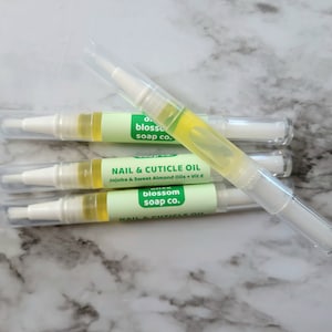 May include: Four nail and cuticle oil pens with clear caps. Each pen has a white applicator tip and a light green label with the text "Nail & Cuticle Oil" and "Jojoba & Sweet Almond Oils + Vit E". The oil is yellow.