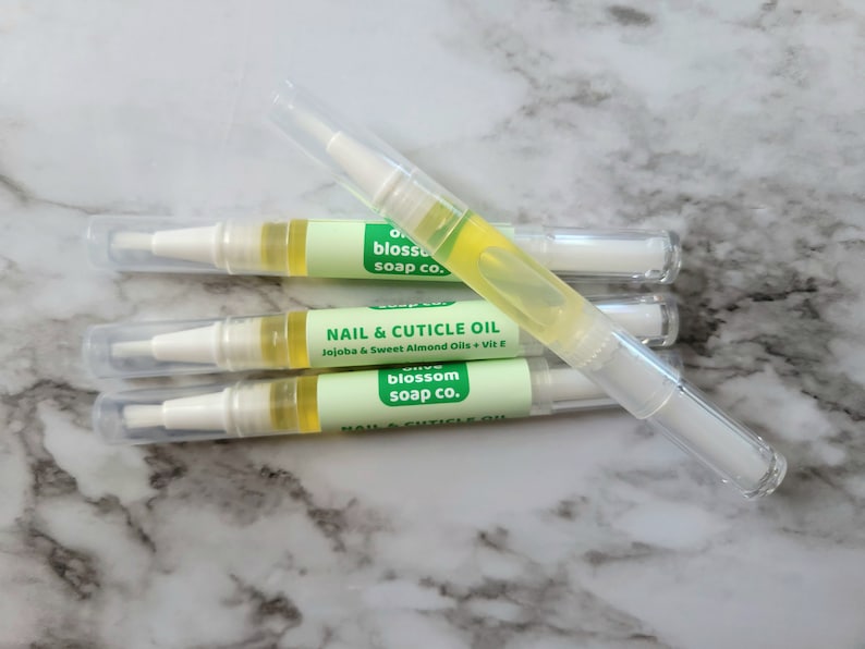 May include: Four nail and cuticle oil pens with clear caps. Each pen has a white applicator tip and a light green label with the text "Nail & Cuticle Oil" and "Jojoba & Sweet Almond Oils + Vit E". The oil is yellow.