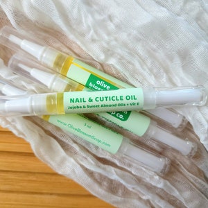 May include: Several nail and cuticle oil pens are arranged on a light-colored surface. Each pen has a clear plastic tube with a white applicator tip and a green label that reads "NAIL & CUTICLE OIL" and "Jojoba & Sweet Almond Oils + Vit E".