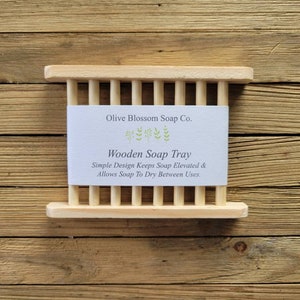 May include: A light-colored wooden soap tray with a slatted design. The tray has a rectangular shape with vertical wooden slats. A white label with the text "Olive Blossom Soap Co." and "Wooden Soap Tray" is centered on the tray.