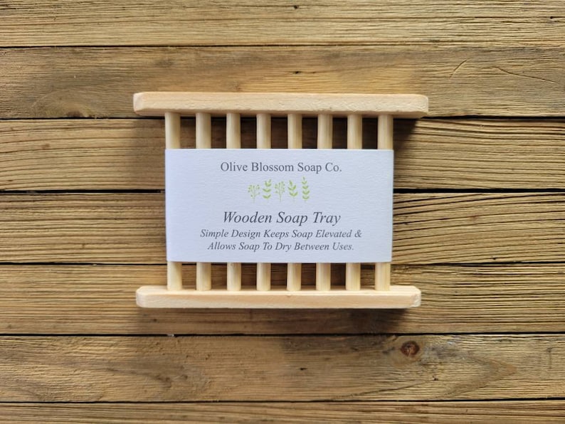 May include: A light-colored wooden soap tray with a slatted design. The tray has a rectangular shape with vertical wooden slats. A white label with the text "Olive Blossom Soap Co." and "Wooden Soap Tray" is centered on the tray.