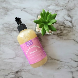 May include: A clear plastic bottle with a black pump dispenser containing yellow liquid hand soap. The pink label reads olive blossom soap co. BLACK RASPBERRY VANILLA Moisturizing Hand Soap Olive Oil + Dead Sea Salt 8 Fl Oz.