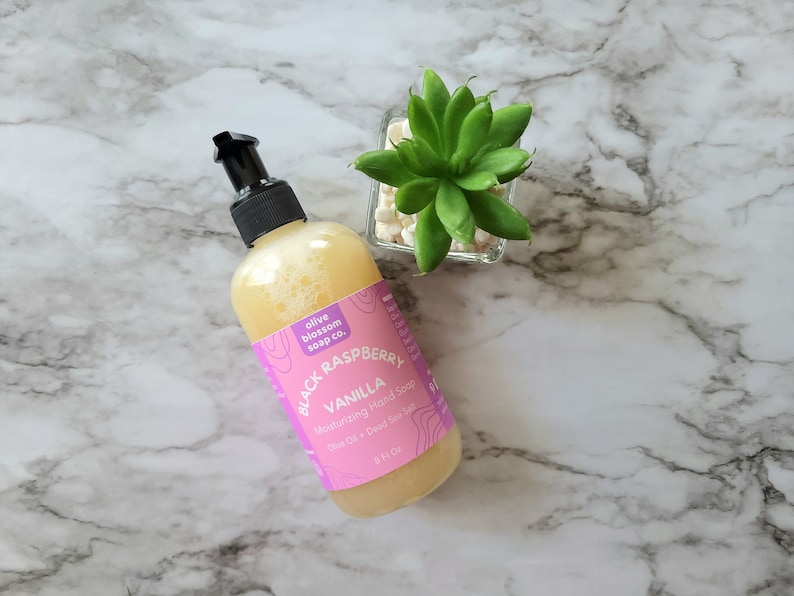 May include: A clear plastic bottle with a black pump dispenser containing yellow liquid hand soap. The pink label reads olive blossom soap co. BLACK RASPBERRY VANILLA Moisturizing Hand Soap Olive Oil + Dead Sea Salt 8 Fl Oz.