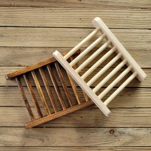 May include: Two wooden soap dishes. One is stained a dark brown, the other is natural wood. Each has a rectangular frame with evenly spaced wooden slats. The dishes are angled on a wooden surface.