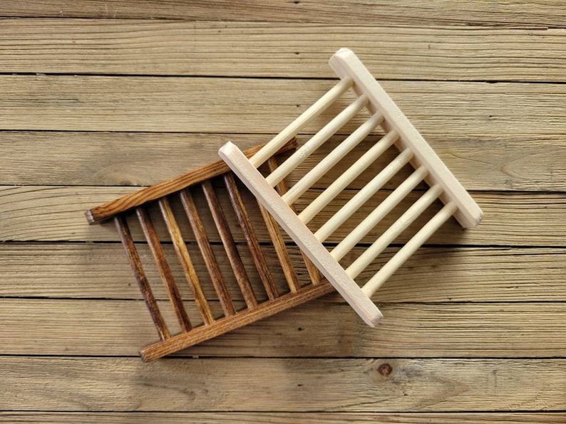 May include: Two wooden soap dishes. One is stained a dark brown, the other is natural wood. Each has a rectangular frame with evenly spaced wooden slats. The dishes are angled on a wooden surface.