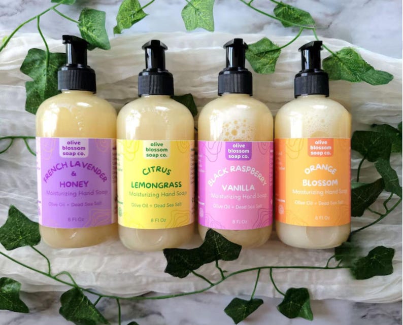May include: Four clear plastic bottles of moisturizing hand soap with black pump dispensers. Each bottle features a colorful label with the text Olive Blossom Soap Co. and scents: French Lavender & Honey, Citrus Lemongrass, Black Raspberry Vanilla, and Orange Blossom, each containing 8 fl oz.