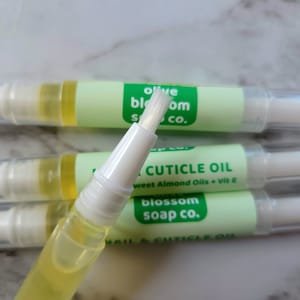 May include: Close-up of several nail and cuticle oil pens. Each pen has a clear tube filled with yellow oil, a white applicator brush, and a light green label with the text "olive blossom soap co." and "NAIL & CUTICLE OIL".