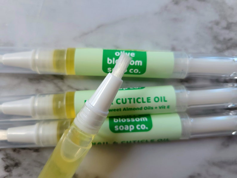 May include: Close-up of several nail and cuticle oil pens. Each pen has a clear tube filled with yellow oil, a white applicator brush, and a light green label with the text "olive blossom soap co." and "NAIL & CUTICLE OIL".
